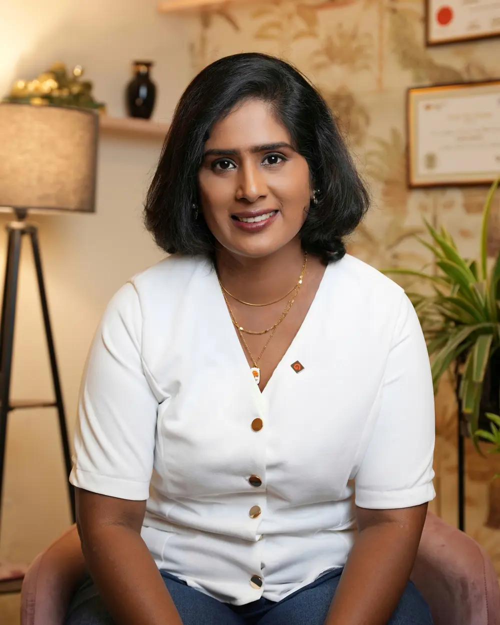 Dr. Indhu Nagaiah in a white blouse, seated and smiling in a warmly lit room with framed certificates