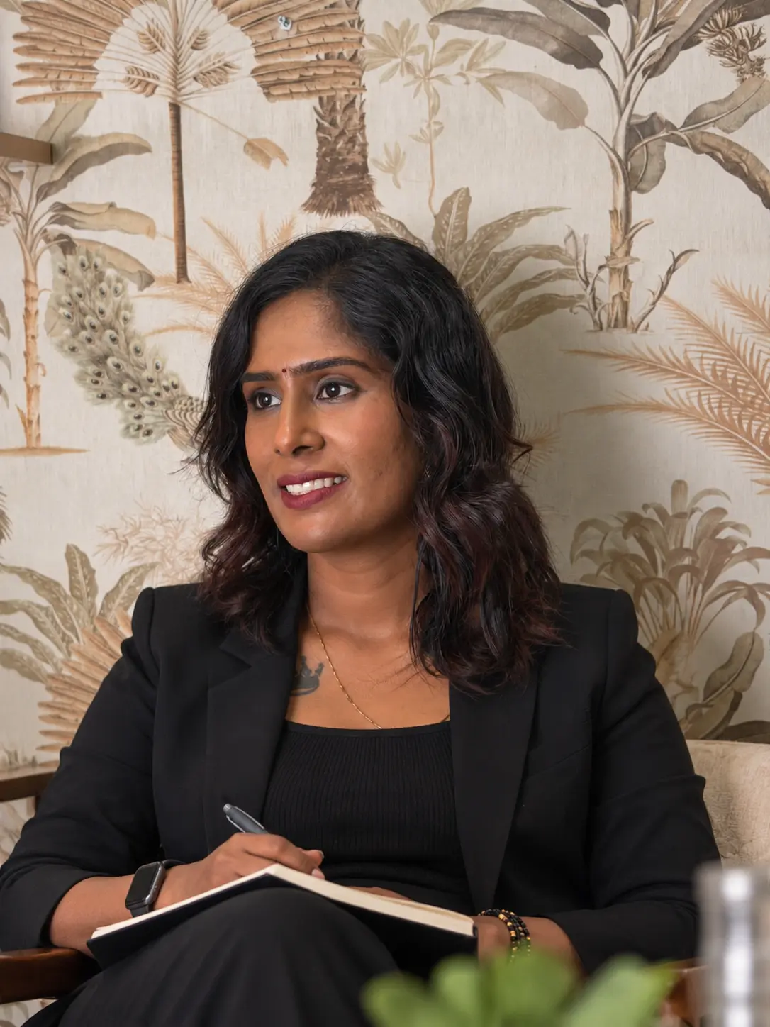 Dr. Indhu Nagaiah in a black blazer, smiling and listening with a notebook in hand, in front of botanical wallpaper