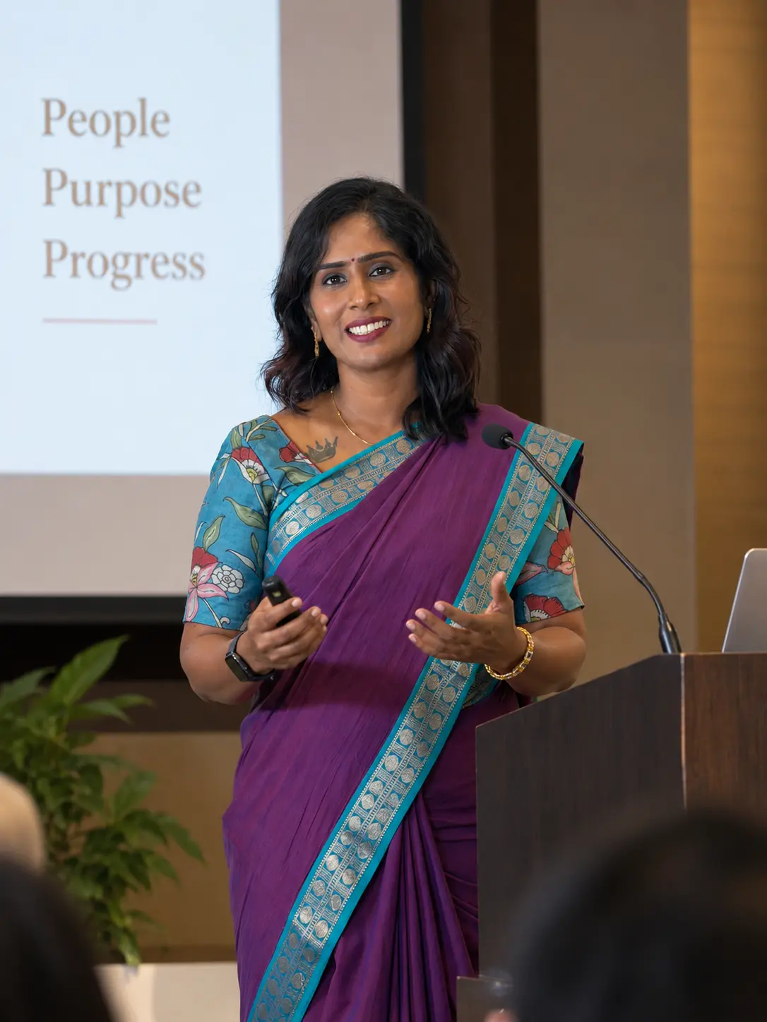 Dr. Indhu Nagaiah in a purple saree, smiling and gesturing as she speaks from a podium